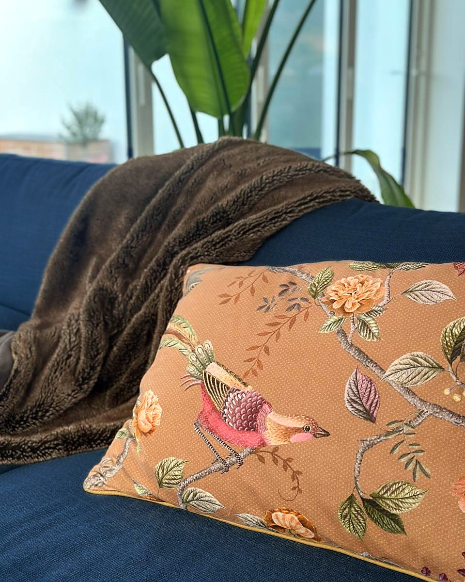 Brunt plaid og pude med fugle og blomster i okker på en mørkeblå sofa foran et vindue
